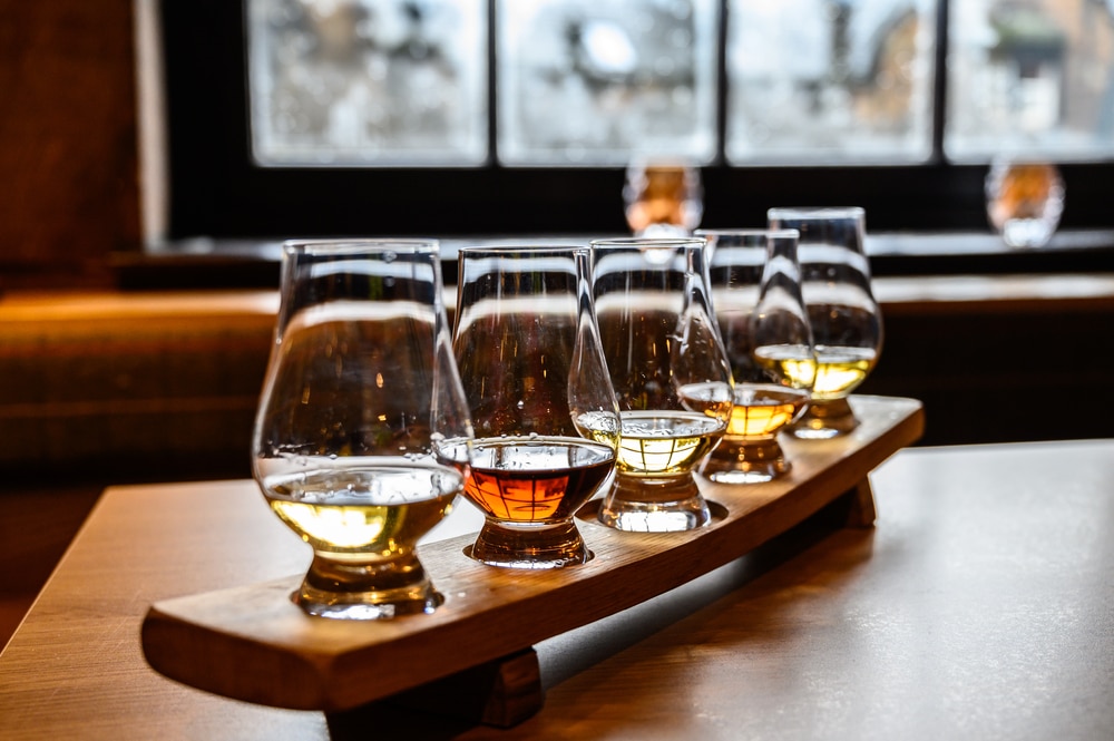 A wooden tray holds five glasses of whiskey in varying shades of amber, arranged in a row on a table, with a blurred window and cozy indoor setting in the background.