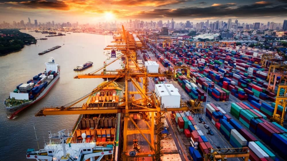 A busy port at sunset with large yellow cranes, shipping containers in various colors, docked cargo ships, and a city skyline in the background.