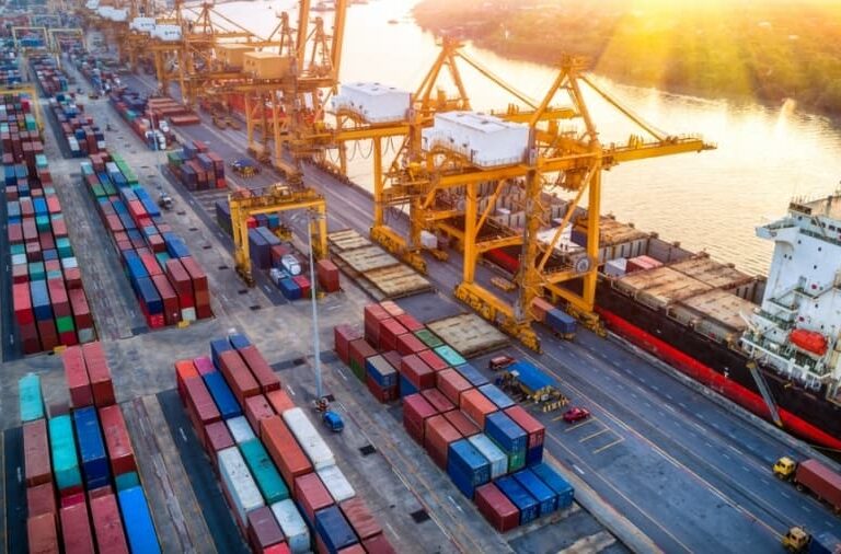 Aerial view of a busy 3PL shipping port with large yellow cranes, stacked colorful shipping containers, and a cargo ship docked by the water at sunset.