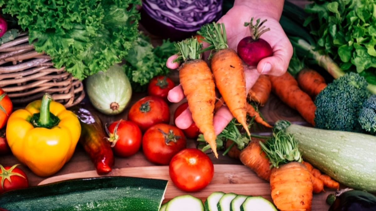 A variety of fresh vegetables, including carrots, tomatoes, bell peppers, lettuce, radishes, broccoli, zucchini, and cabbage—all handled in compliance with FSMA guidelines—are spread out on a wooden surface. A hand holds up three carrots and a radish.