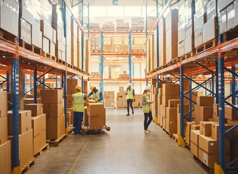 Warehouse workers in safety vests organize and move large cardboard boxes along wide aisles lined with tall metal shelves, efficiently tracking inventory with a WMS. Bright lighting and high ceilings are visible.
