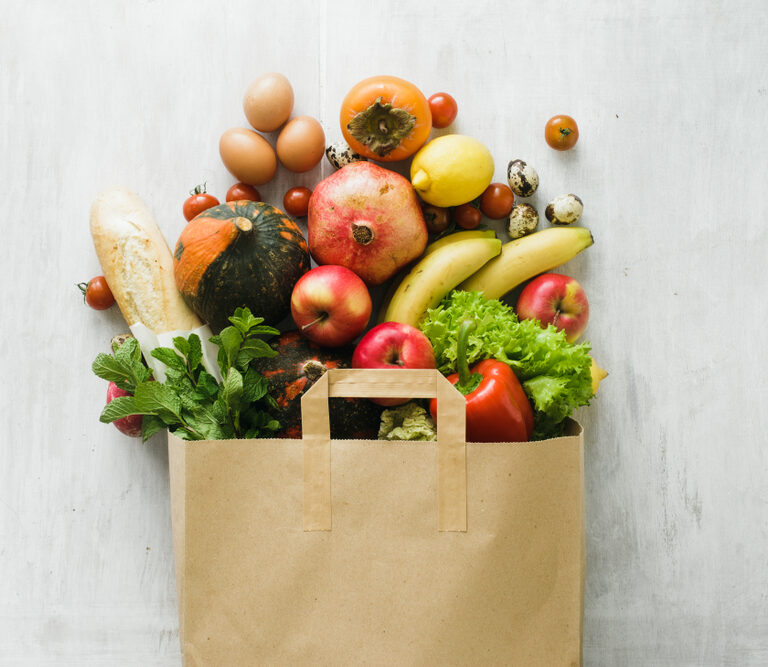 A brown paper bag overflowing with fresh groceries, including eggs, bread, pumpkins, apples, bananas, lettuce, tomatoes, a pomegranate, lemons, strawberries, mint, and quail eggs on a white surface—perfect for FSMA-compliant kitchens.