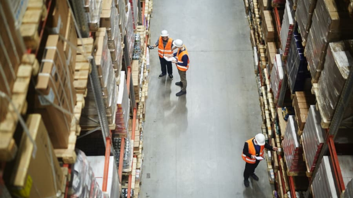 Overhead view of three workers in orange safety vests and hard hats walking and inspecting shelves in a large warehouse filled with stacked boxes and packages.