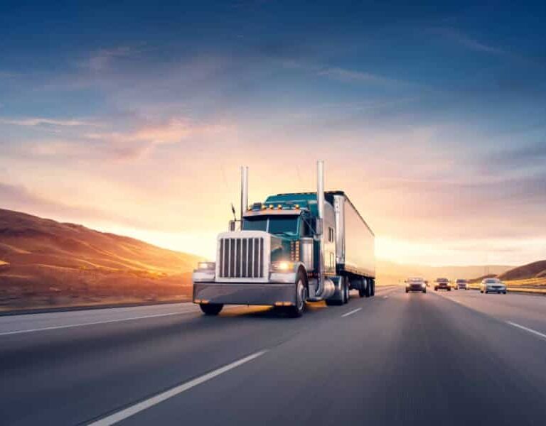 A large semi-truck drives down a multi-lane highway at sunset, with a few cars visible in adjacent lanes and rolling hills in the background under a dramatic sky.