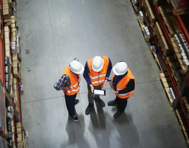 Three people wearing white hard hats and orange safety vests stand in a warehouse aisle, looking at a tablet. Shelves stocked with boxes and items are visible on both sides.