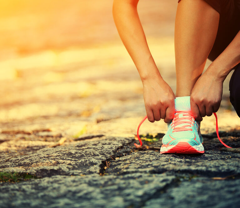 A person tying the laces of a bright teal and pink running shoe while crouching on a sunlit stone path, preparing for a run or workout.
