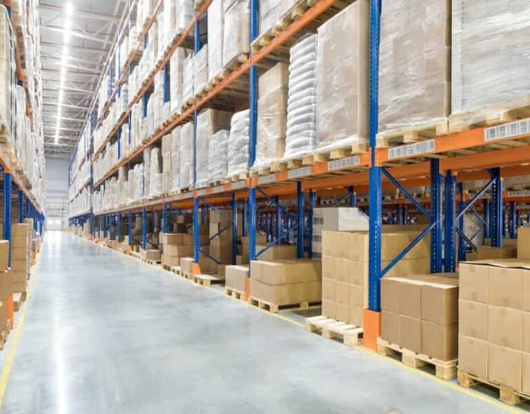 Large warehouse interior with tall blue and orange industrial shelving, stacked with cardboard boxes and wrapped pallets; the polished concrete floor and wide aisle highlight a space designed for efficient warehouse automation.