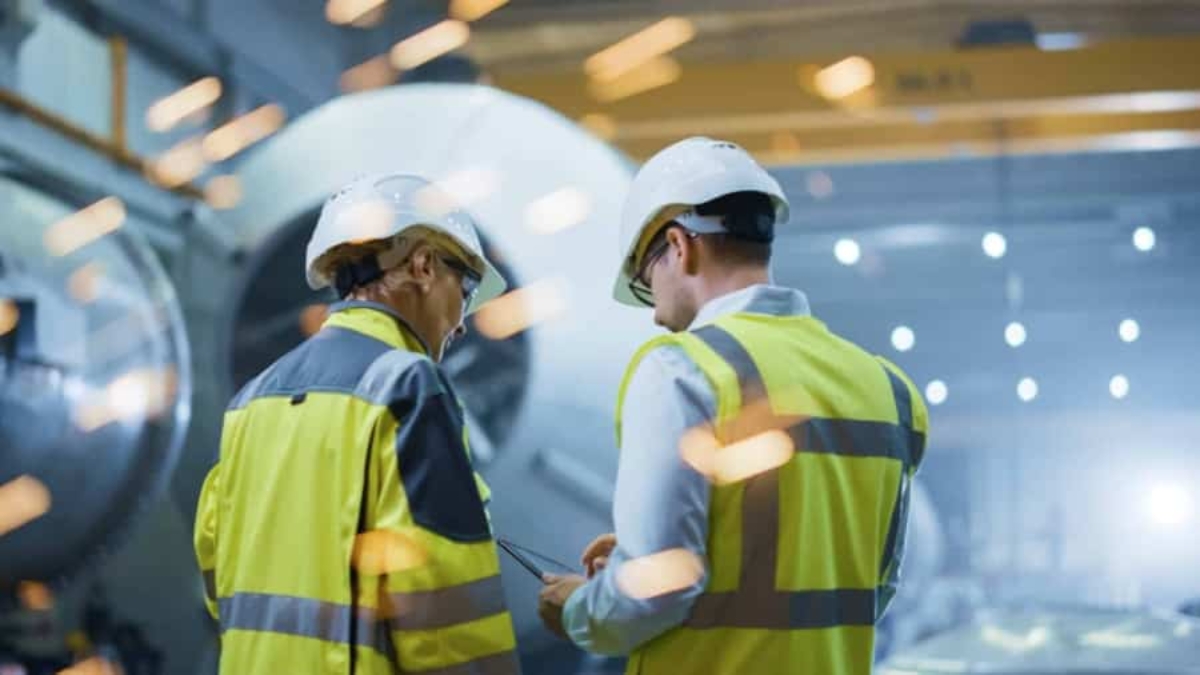 Two workers wearing safety vests and helmets stand in an industrial setting, with machinery and bright flying sparks in the background, suggesting active manufacturing or metalworking.