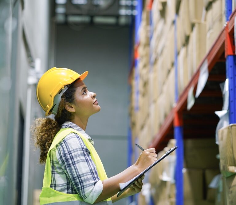 A woman wearing a yellow hard hat and safety vest stands in a warehouse, promoting warehouse safety as she holds a clipboard and pen, looking up at shelves stacked with cardboard boxes.