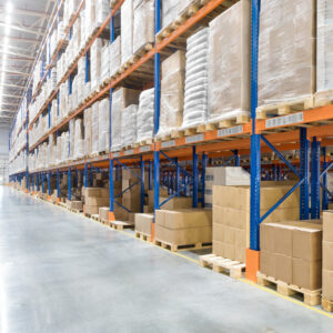 Rows of tall warehouse shelves filled with stacked cardboard boxes and pallets line a wide, clean aisle in a brightly lit industrial storage facility.