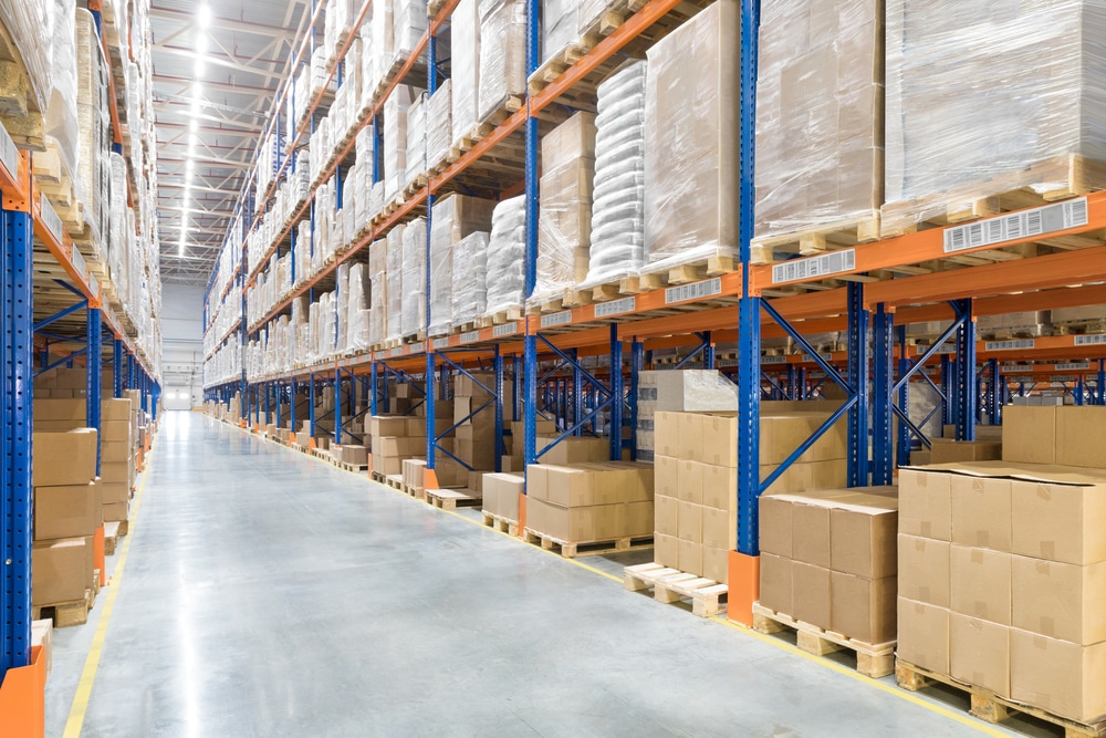 Rows of tall warehouse shelves filled with stacked cardboard boxes and pallets line a wide, clean aisle in a brightly lit industrial storage facility.