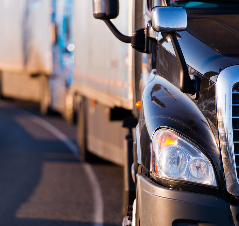 A close-up of the front of a black semi-truck driving on a road, with several other trucks in a line behind it—all traveling in the same direction and highlighting the importance of cyber-attacks prevention in transportation.