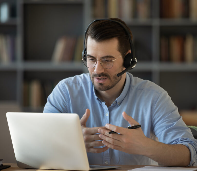 A man wearing glasses and a headset sits at a desk in front of a laptop, gesturing with his hands while speaking during a video call about Microsoft’s Sales Copilot. Bookshelves are visible in the background.