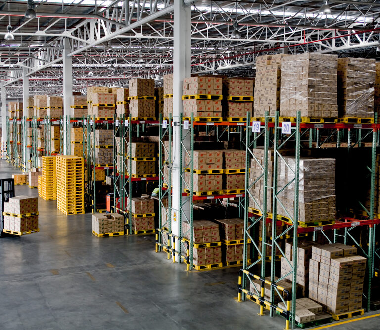 A large warehouse interior with tall shelves stacked with boxes and pallets, featuring efficient slotting and a worker driving a forklift along the aisle. The space is well-lit and organized, with industrial shelving and high ceilings.