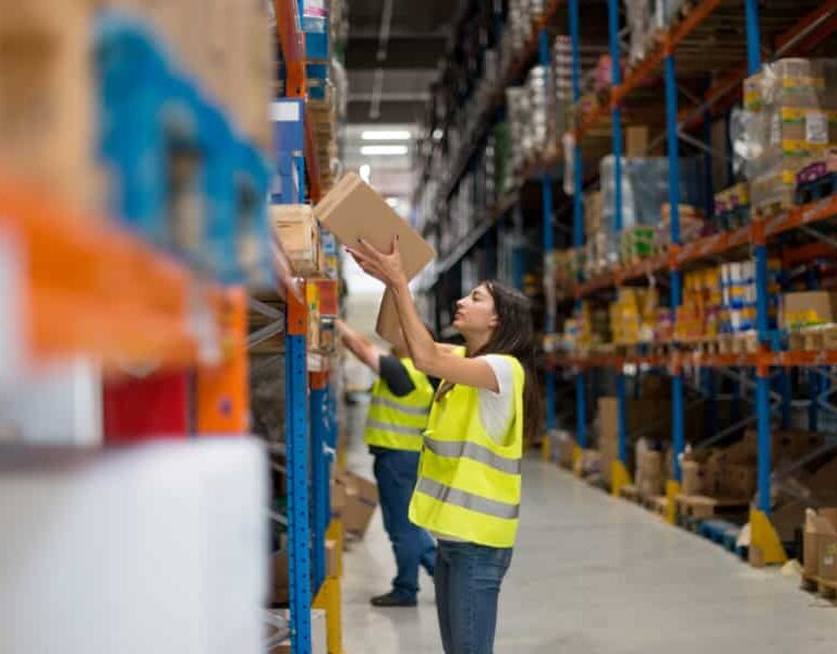 A woman in a yellow safety vest places a box on a high shelf in a warehouse, while another worker in a vest organizes items on a nearby shelf. Shelves are filled with various boxes and products.