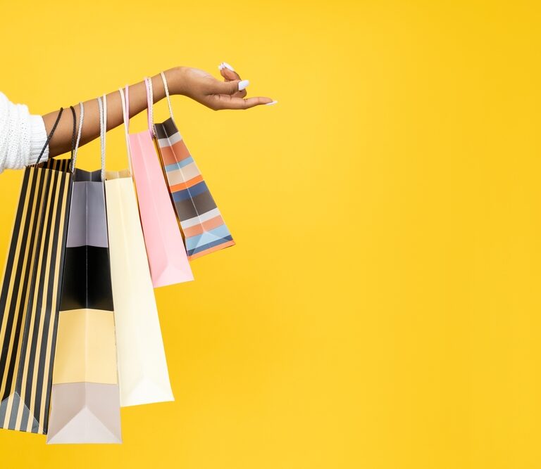 A persons outstretched arm holds several colorful shopping bags against a bright yellow background. The bags have various patterns and colors, including stripes and solid tones.