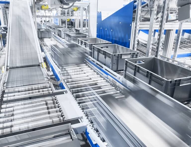 A modern automated conveyor system transports black plastic bins along metal rollers in a bright, industrial warehouse setting. The machinery and containers are in motion, suggesting efficient, high-speed operations.