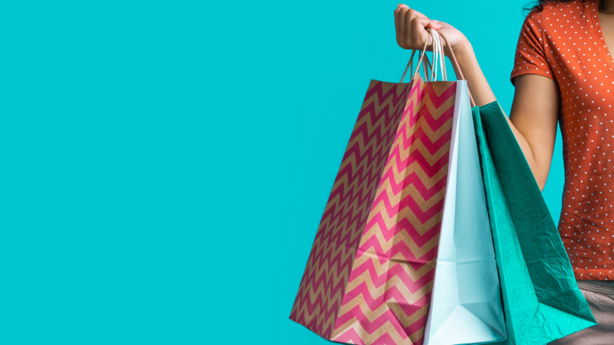 A person wearing an orange polka dot shirt holds colorful shopping bags against a bright turquoise background. Only the persons arm and part of their torso are visible.