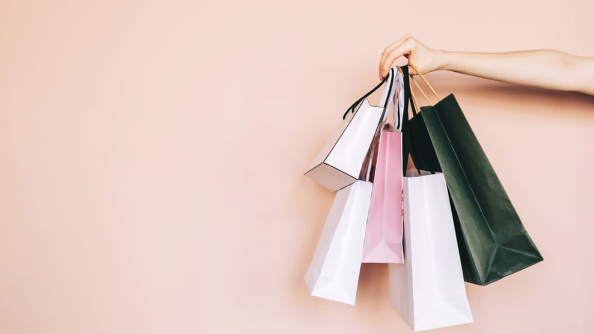 A hand is holding several colorful shopping bags by their handles against a light peach background, highlighting the variety that WMS can help organize. The bags vary in size and color, including white, pink, black, and dark green.