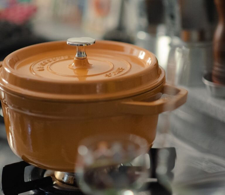 An orange dutch oven with a silver knob sits on a gas stovetop in a kitchen, with blurred kitchen items and utensils visible in the background.