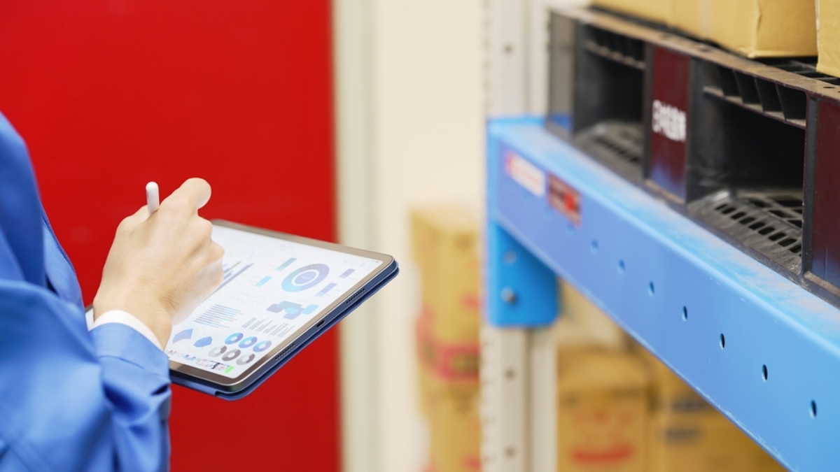 Person in blue uniform holds a tablet displaying WMS charts and graphs while standing near warehouse shelves with boxes.