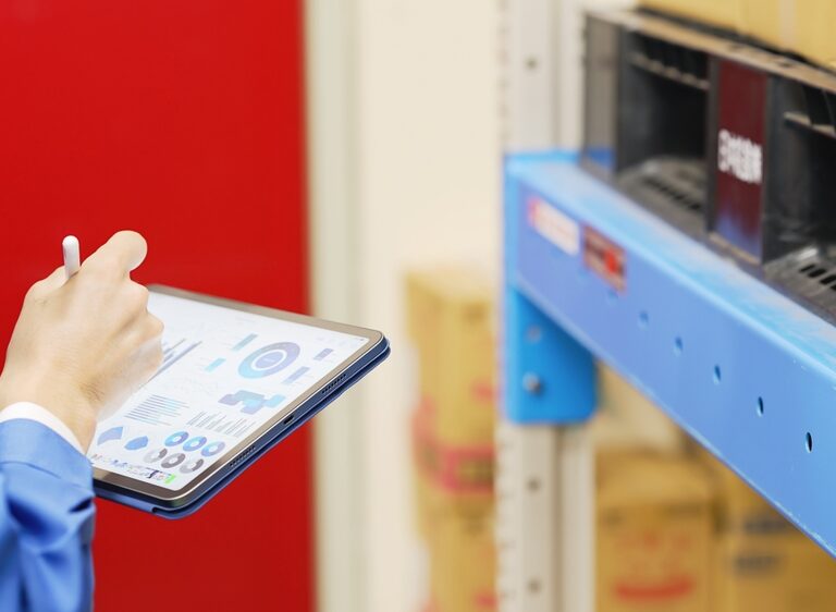 Person in blue uniform holds a tablet displaying WMS charts and graphs while standing near warehouse shelves with boxes.