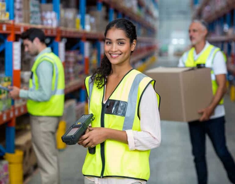 A smiling woman in a yellow safety vest holds a barcode scanner in a warehouse, with two coworkers working in the background among shelves stocked with boxes and goods.