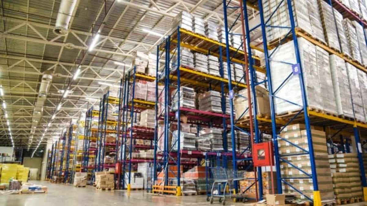 Large warehouse interior with tall blue and yellow racks stacked with pallets of boxes and goods. Wide aisles separate the shelves, and bright lighting illuminates the organized storage area.