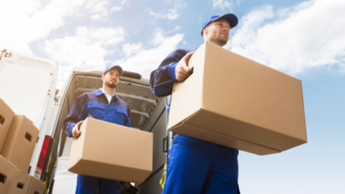 Two movers wearing blue uniforms carry cardboard boxes from the back of a white van under a bright blue sky with scattered clouds. More boxes are stacked nearby.