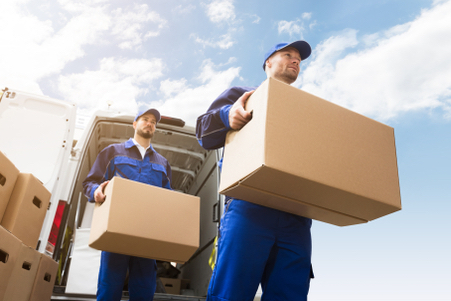 Two movers wearing blue uniforms carry cardboard boxes from the back of a white van under a bright blue sky with scattered clouds. More boxes are stacked nearby.