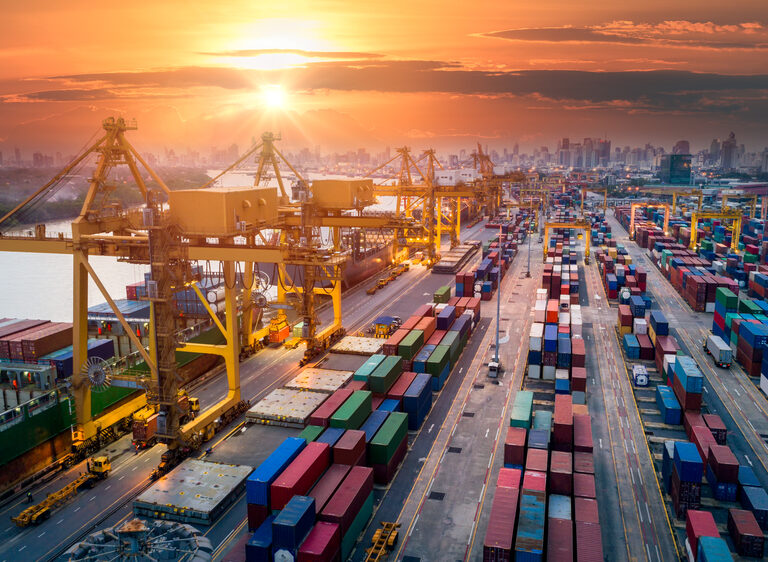 Aerial view of a busy shipping port at sunset, with large yellow cranes, colorful shipping containers, and a city skyline in the background. The sun is setting over the horizon, casting a warm glow on the scene.