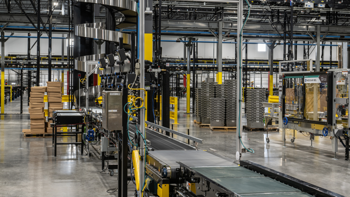 Busy warehouse interior with conveyor belts, stacked cardboard boxes, metal shelves, automated machinery, and organized workstations, powered by SLAM solutions for an industrial and highly automated environment.