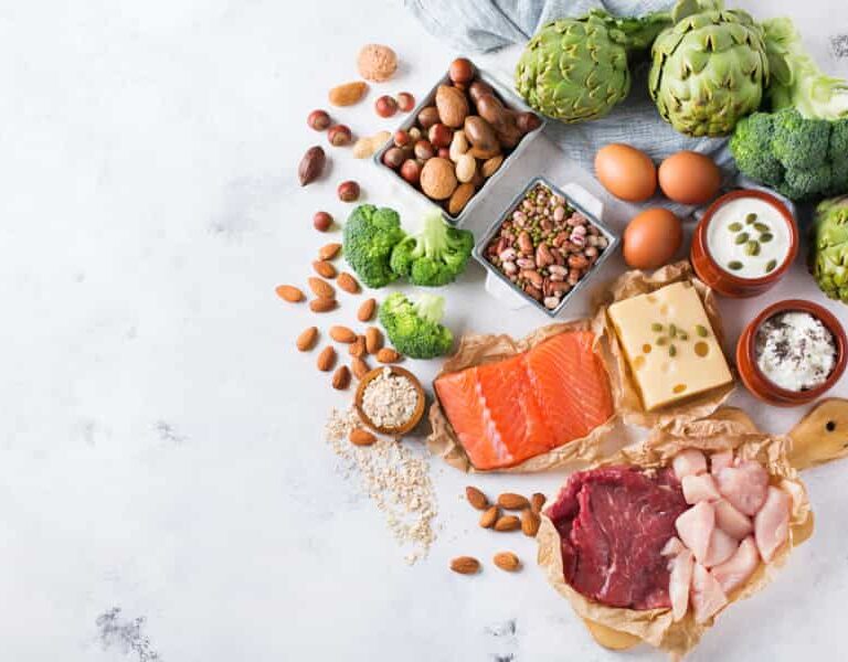 Assorted healthy foods including salmon, beef, chicken, cheese, eggs, nuts, seeds, yogurt, artichokes, broccoli, and beans arranged on a white surface.