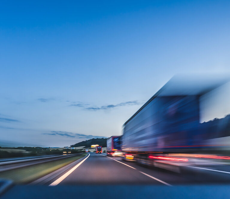 A blurred truck speeds down a highway at dusk, creating motion blur. The sky is clear with soft blue tones, and the distant landscape is faintly visible beside the road—capturing the essence of modern transportation sourcing in motion.