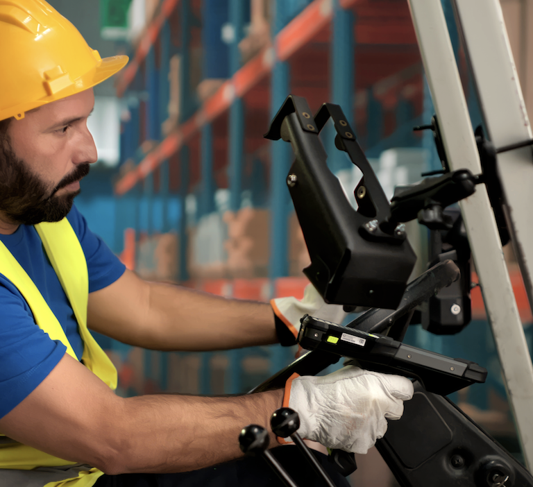 A warehouse worker wearing a yellow hard hat, safety vest, and gloves operates a forklift in a storage area with shelves filled with boxes, efficiently managing inventory using an ERP system.