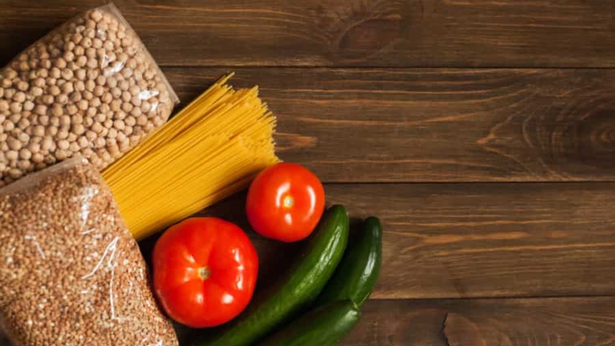 A flat lay of two tomatoes, two cucumbers, uncooked spaghetti, dried chickpeas, and grains on a dark wooden surface.