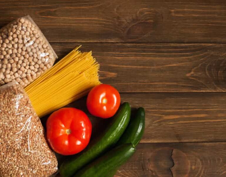 A flat lay of two tomatoes, two cucumbers, uncooked spaghetti, dried chickpeas, and grains on a dark wooden surface.