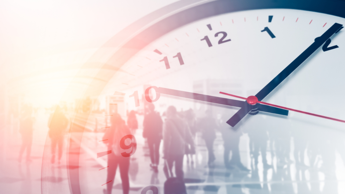 A close-up of a clock face showing the time as 10:09, with a blurred background of people walking in a brightly lit indoor space, creating a sense of motion and passing time.