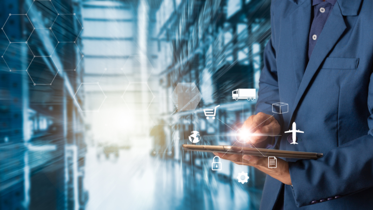 A person in a suit uses a tablet, with logistics and supply chain icons like a truck, cart, box, airplane, and lock overlaid. The background shows a blurred warehouse or industrial setting.
