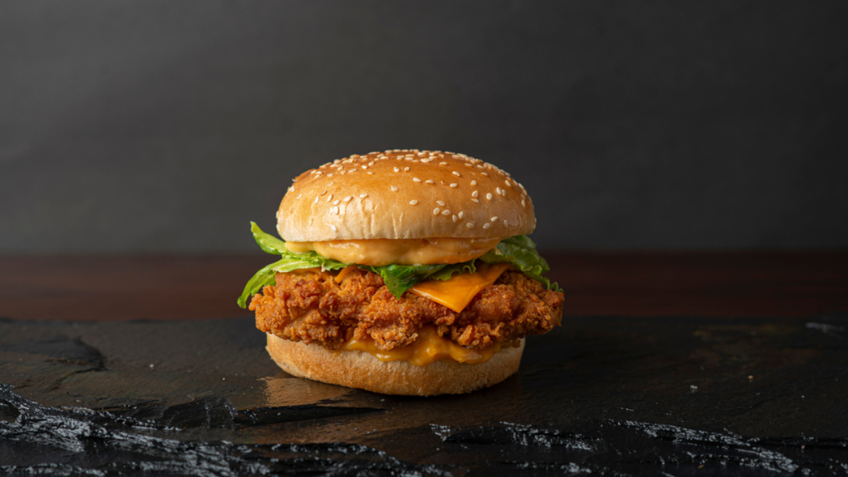 A crispy fried chicken sandwich with lettuce, cheddar cheese, and sauce on a sesame seed bun, placed on a dark slate surface with a plain dark background.