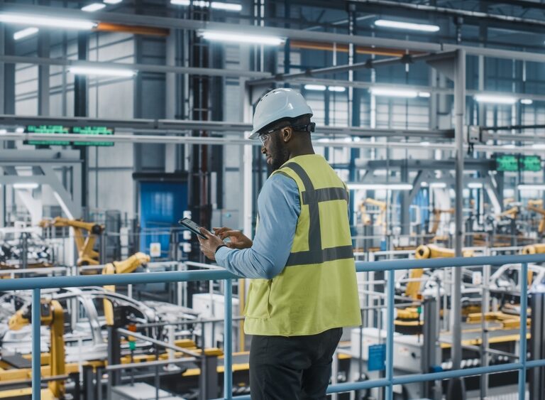 A person wearing a white hard hat and yellow safety vest uses a tablet while standing in a modern, automated factory with robotic arms and machinery in the background.