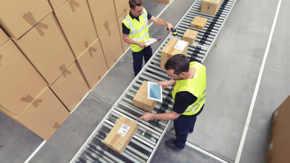 Two warehouse workers in yellow safety vests process parcels on a conveyor belt, scanning and checking boxes. Stacks of cardboard boxes are arranged along the sides of the conveyor in a large, organized warehouse space.