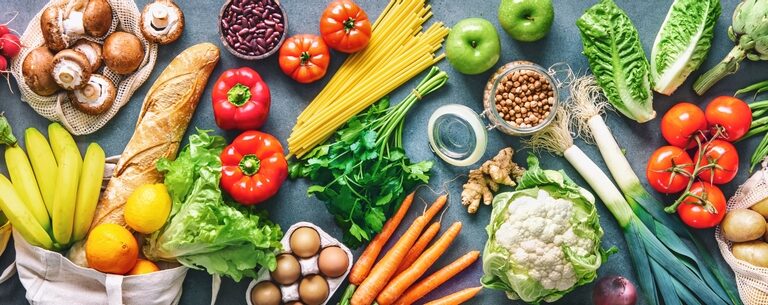 A variety of fresh foods including vegetables, fruits, bread, eggs, pasta, beans, olive oil, and milk are arranged on a table, showcasing healthy ingredients for balanced meals.