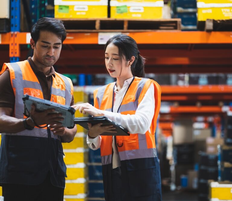 Two warehouse workers wearing orange safety vests review labor management information on tablets while standing in an aisle filled with shelves of yellow and blue storage bins.