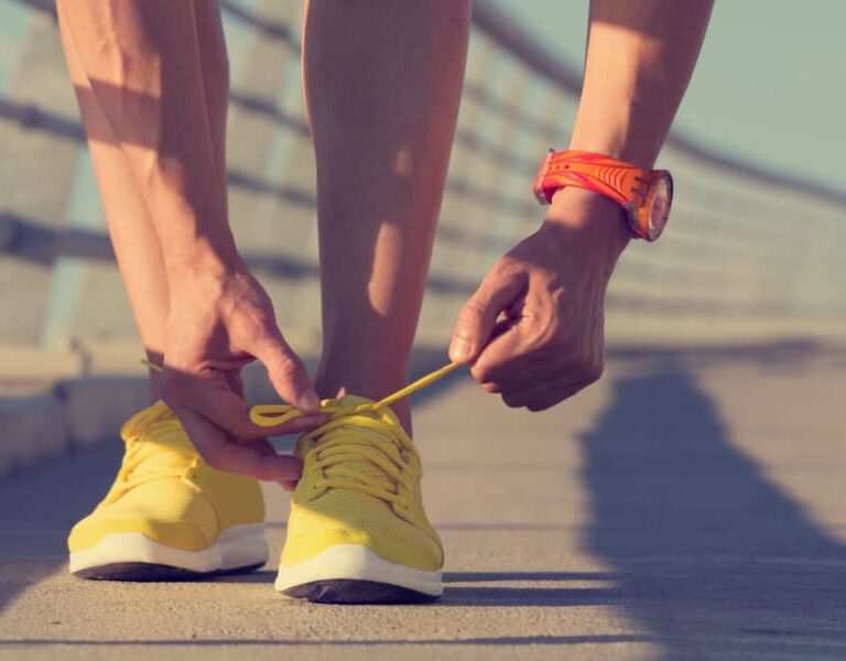 A person bends down to tie the yellow shoelaces on their bright yellow running shoes while standing on a sunlit outdoor pathway, wearing an orange watch on their wrist.