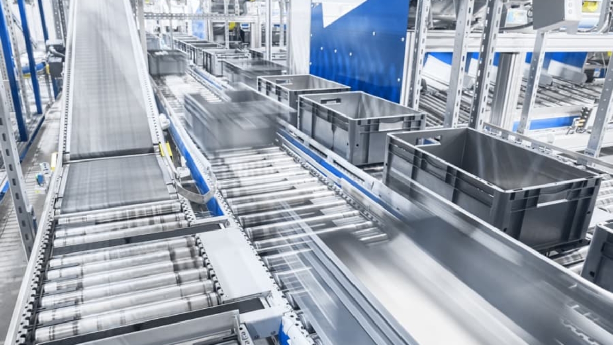 Automated conveyor system with gray plastic bins moving quickly along metal rollers in a bright, modern warehouse or distribution center showcases how facilities automate processes for fast-paced, efficient operations. Motion blur highlights speed and productivity.
