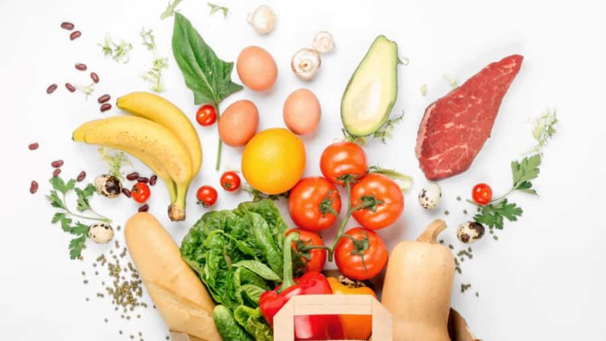 A paper bag overflowing with fresh groceries—including tomatoes, lettuce, bananas, eggs, avocado, steak, and more—on a white background highlights the importance of supply chain traceability for quality and safety.