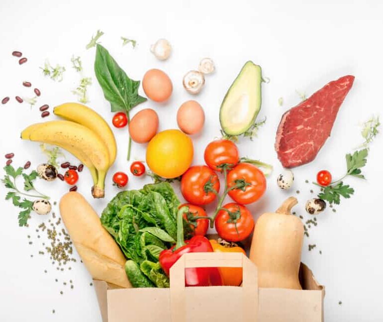 A paper bag overflowing with fresh groceries—including tomatoes, lettuce, bananas, eggs, avocado, steak, and more—on a white background highlights the importance of supply chain traceability for quality and safety.