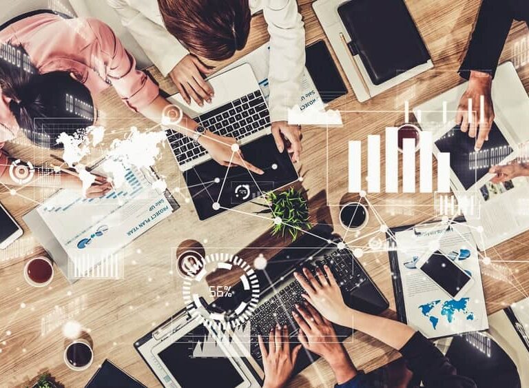 A group of people work together at a table with laptops, tablets, documents, and coffee. Digital graphics overlay the scene, showing charts and data visualizations as they collaborate to select the right ERP system for their business needs.