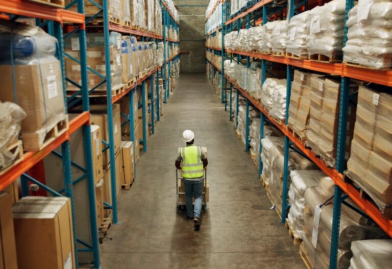 A warehouse worker in a reflective vest and hard hat pushes a cart down a wide aisle between tall shelves filled with stacked boxes and wrapped pallets.